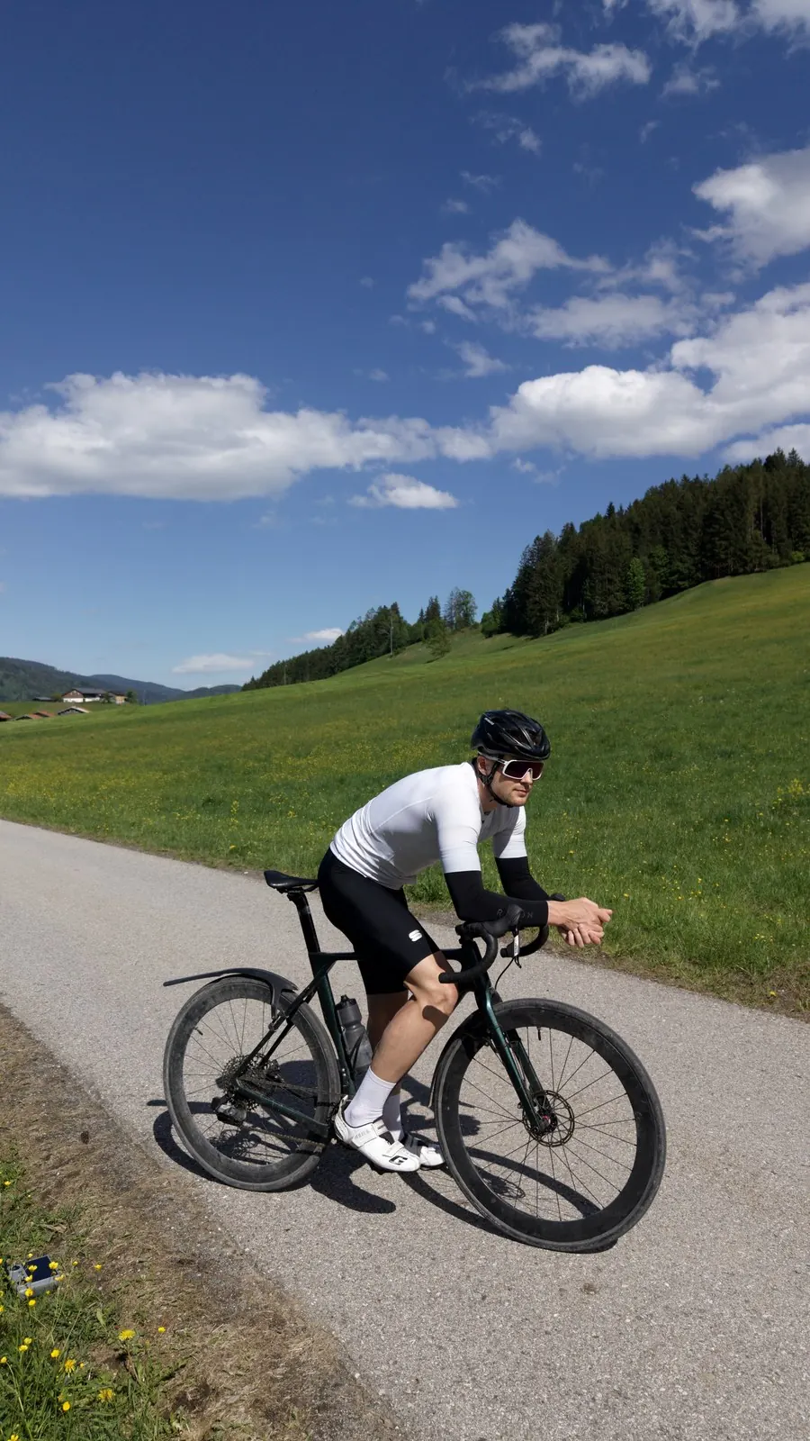 Hans-Jörg Stolz auf dem Rennrad auf einer Straße durch Wiesen, dahinter bewaldete Hänge des Chiemgaus